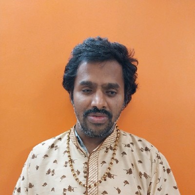 Headshot of Srinivasu in front of an orange wall, wavy black hair and short beard, wearing a cream colored top with a gold pattern and a necklace.