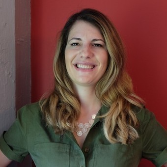 Headshot of Steph standing in front of a red wall, loosely curled brown hair and a green button blouse smiling towards the camera.